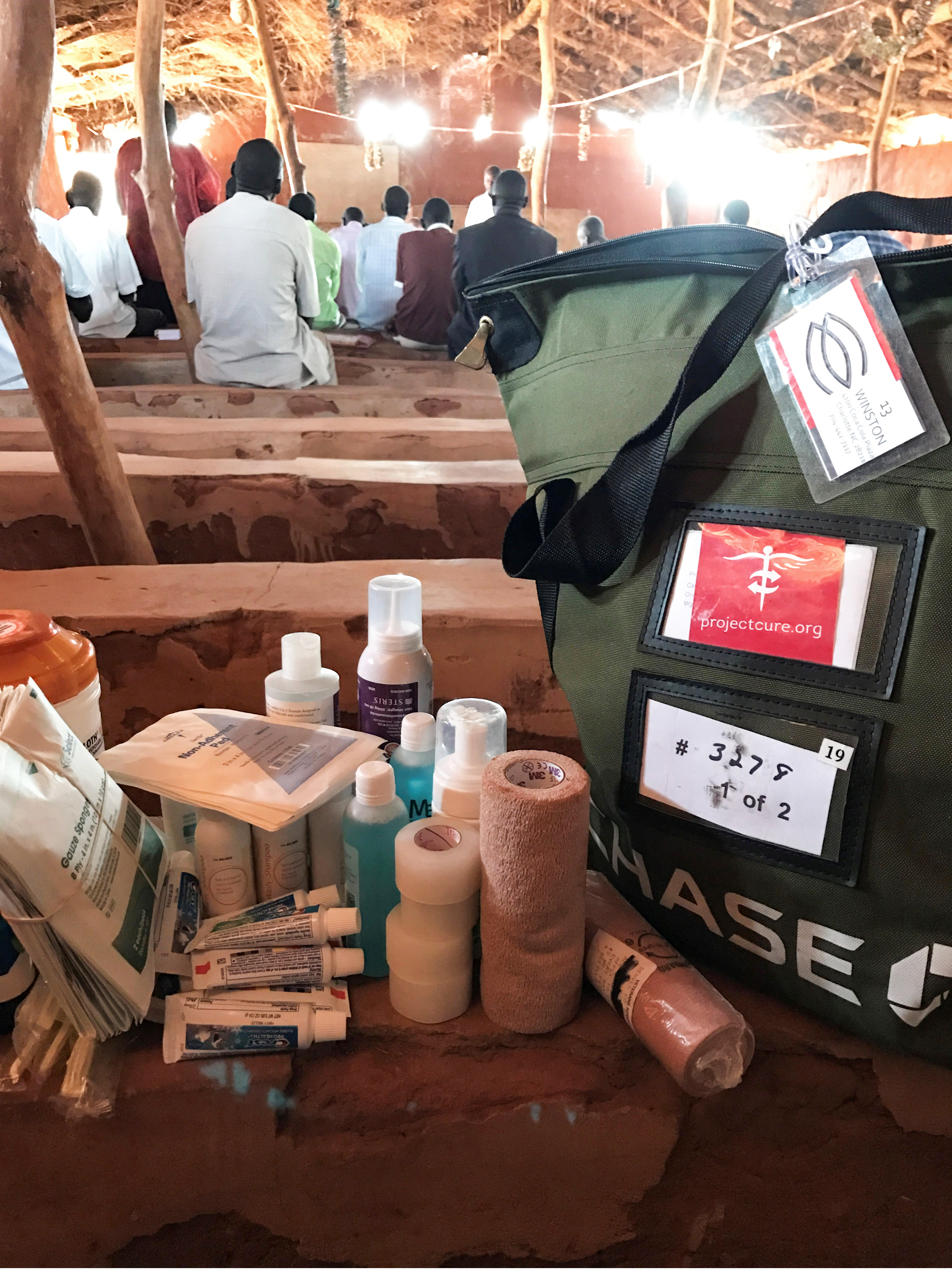 Project C.U.R.E. C.U.R.E. Kit filled with medical supplies including bandages, antiseptics, and hygiene items, placed inside a rural clinic with patients seated in the background in an underserved community.