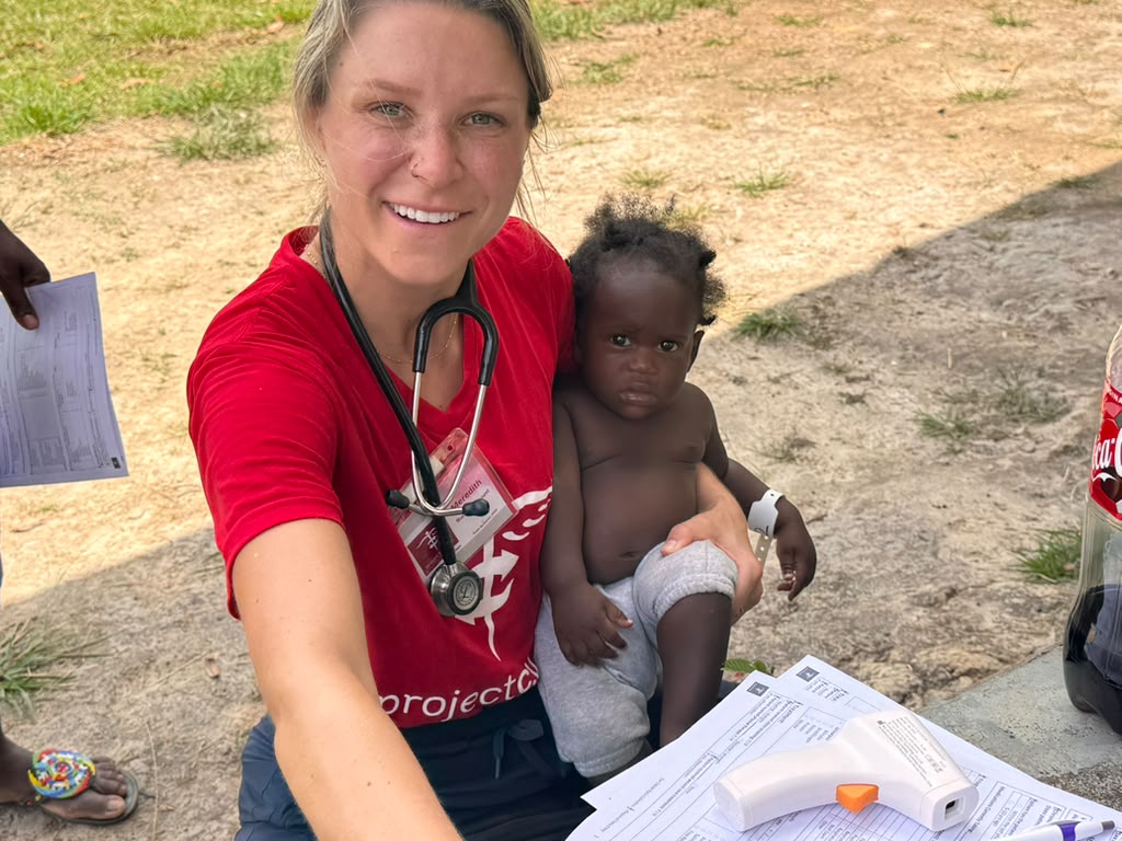 Nursing student Meredith Evilsizer, wearing a red Project C.U.R.E. shirt and stethoscope, holds a young child beside patient intake forms at an outdoor clinic in Suriname.