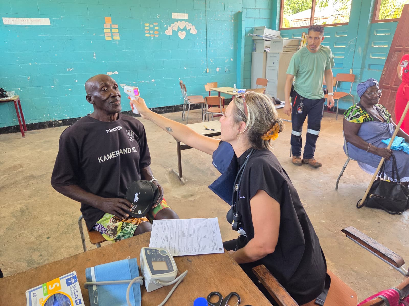 A C.U.R.E. Clinics volunteer takes a patient's temperature with a forehead thermometer inside a turquoise-walled classroom converted into a clinic in Suriname, as other patients wait nearby.